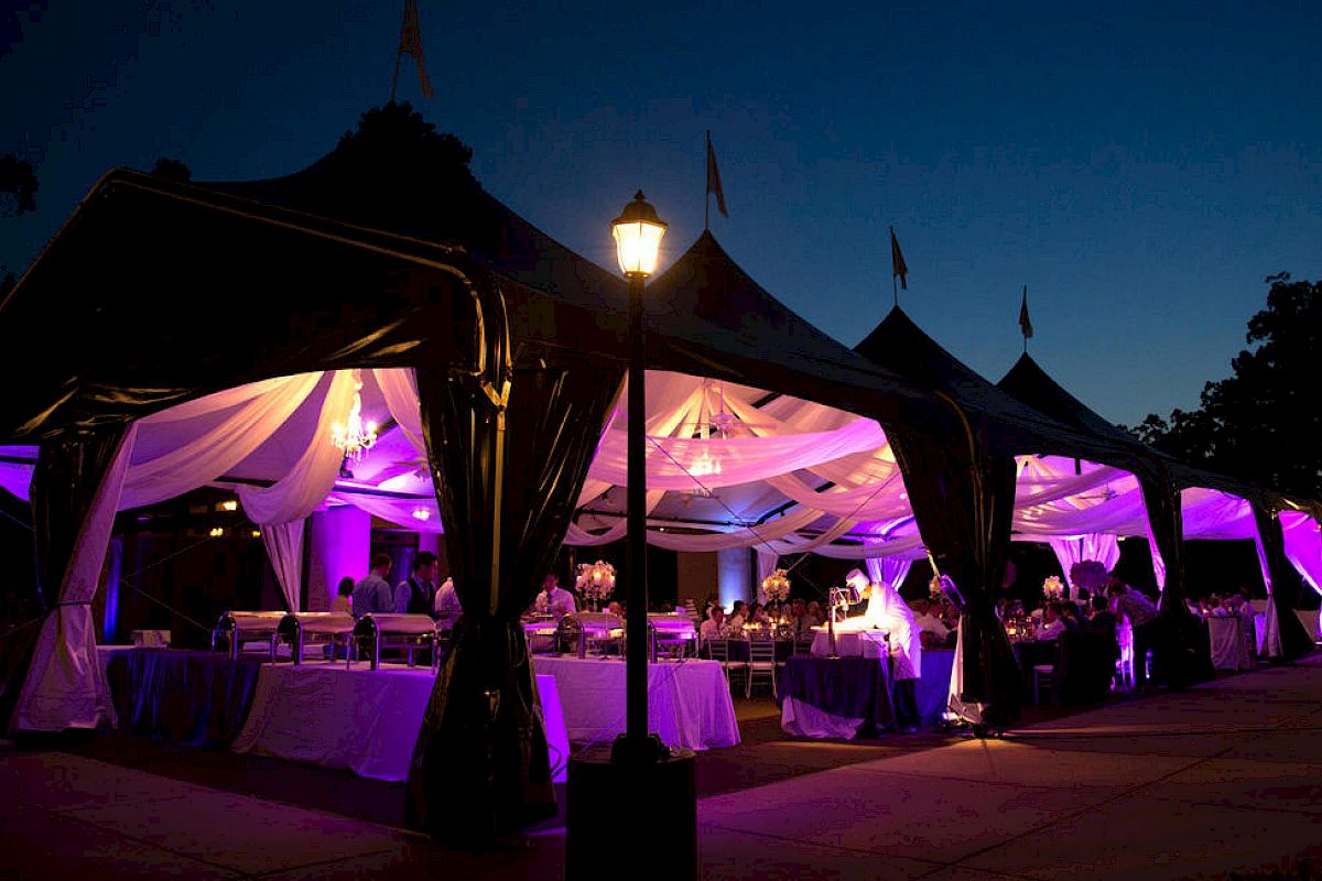 A nighttime outdoor event under elegant, draped tents with purple lighting, banquet tables, and guests dining under warm lanterns.