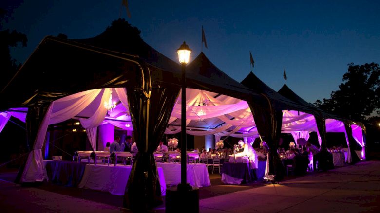 A nighttime outdoor event under elegant, draped tents with purple lighting, banquet tables, and guests dining under warm lanterns.