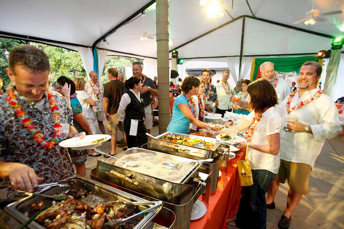 People line up at a buffet under a tent, serving themselves from chafing dishes while others chat and mingle at a casual outdoor party.