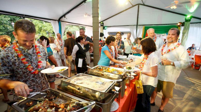 People line up at a buffet under a tent, serving themselves from chafing dishes while others chat and mingle at a casual outdoor party.