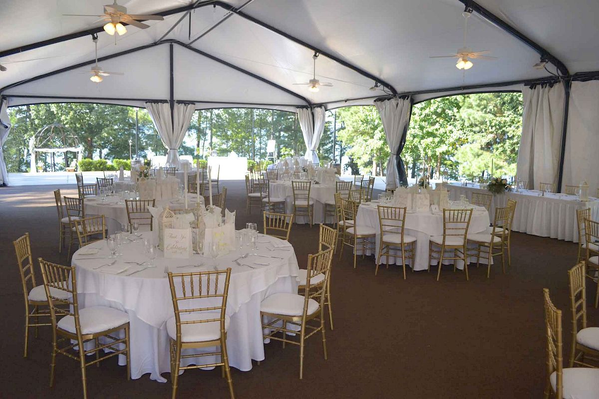 A large event tent set up for a formal reception with round tables, white tablecloths, gold chairs, and soft natural light.