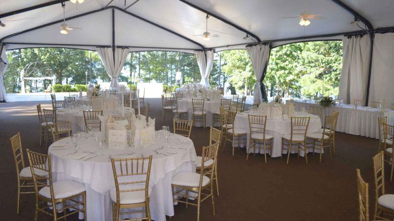 A large event tent set up for a formal reception with round tables, white tablecloths, gold chairs, and soft natural light.