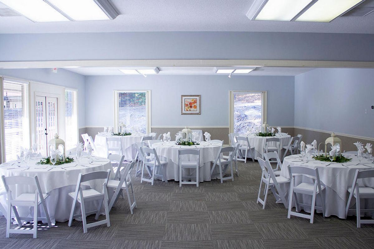 A bright, airy event room with round tables dressed in white tablecloths, white chairs, and simple centerpieces; windows and soft decor.