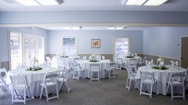 A bright, airy event room with round tables dressed in white tablecloths, white chairs, and simple centerpieces; windows and soft decor.