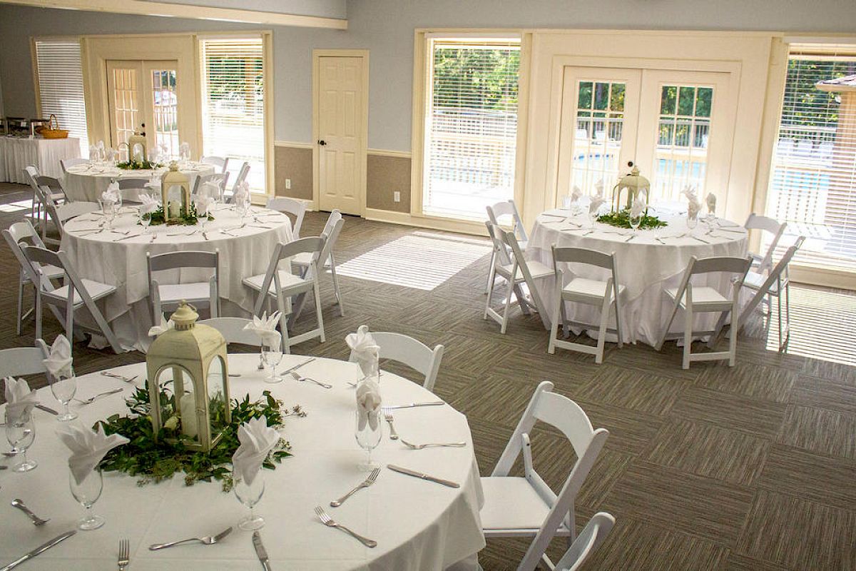 A bright event room set for a formal reception with round tables, white linens, elegant place settings, centerpieces, and lanterns by large windows.