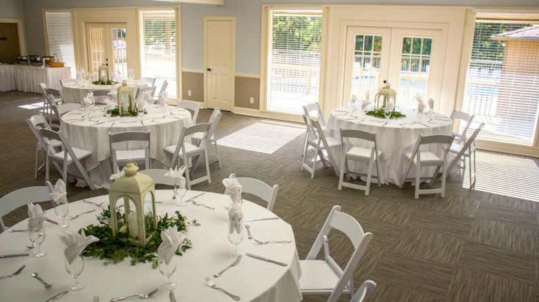 A bright event room set for a formal reception with round tables, white linens, elegant place settings, centerpieces, and lanterns by large windows.