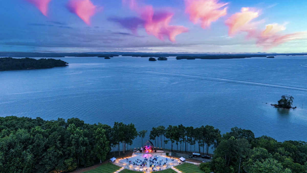 An outdoor event with a stage near a lake at sunset, under pink clouds, surrounded by trees and tents.