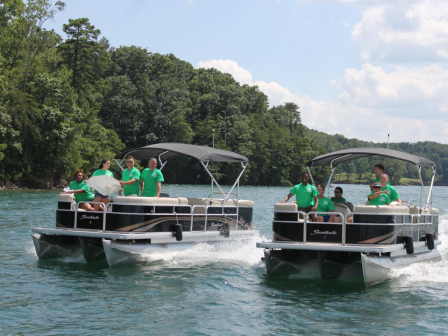 Two pontoon boats with people in green shirts are cruising on a lake, surrounded by trees, under a partly cloudy sky.