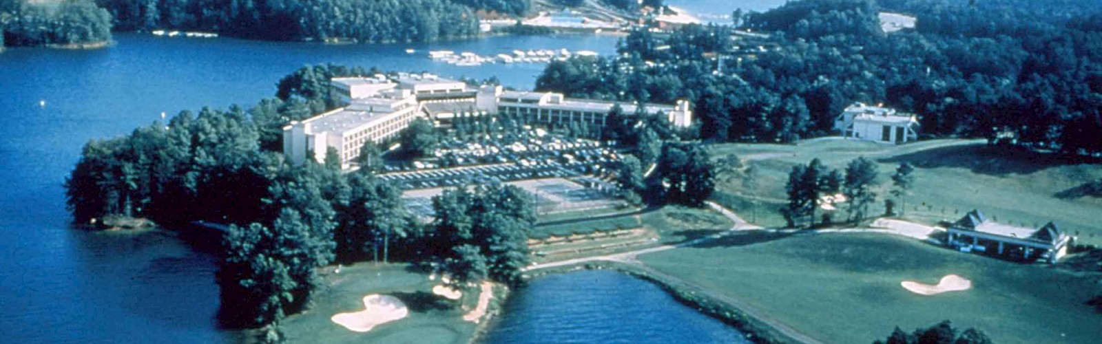 An aerial view of a lakeside resort complex featuring a large building, surrounding wooded areas, a golf course, and multiple water bodies ending the sentence.