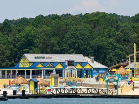 The image shows a colorful beachside or lakeside facility with buildings, lounge areas, and a backdrop of trees.