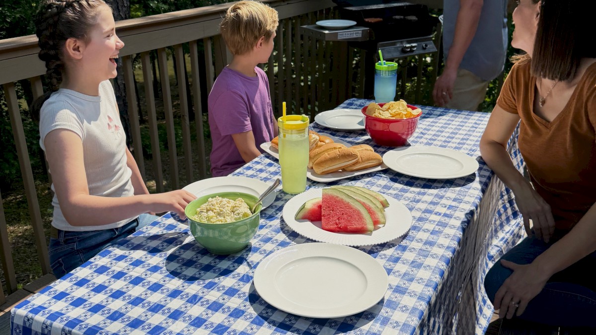 A family is having a barbecue dinner outdoors on a deck, with a table set with food like watermelon, chips, and drinks.