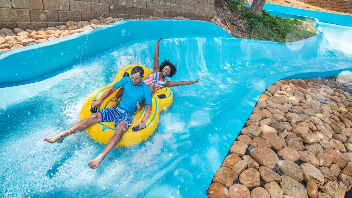 Two people enjoy riding a yellow inflatable tube down a blue water slide outdoors on a sunny day.