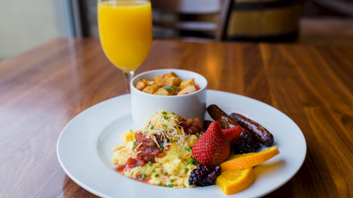 A breakfast plate with scrambled eggs, fresh berries, sausages, and a side of hash browns. A glass of orange juice is in the background.
