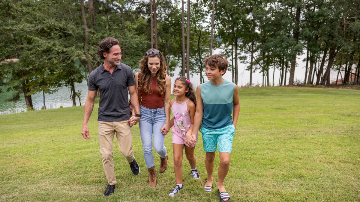 A family of four walks happily through a grassy park near a lake on a cloudy day.