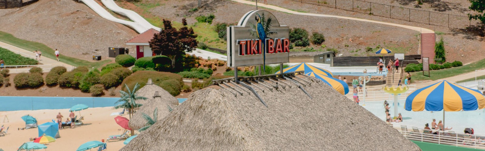 A beach-themed resort with a thatched-roof Tiki Bar, water slides, umbrellas, and several people enjoying the area ending the sentence.