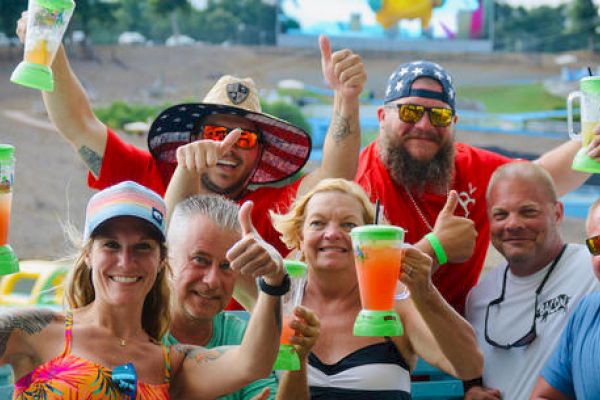 A group of people smiling and holding large drinks, giving thumbs up, outdoors with a background of trees and a hill.