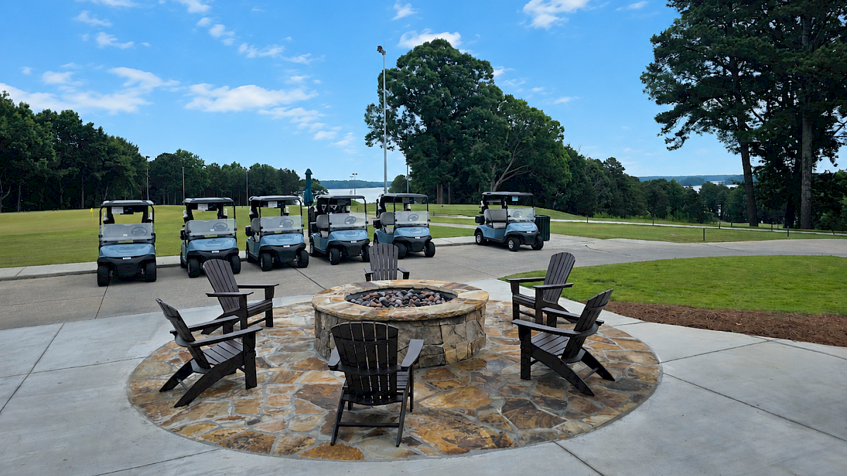 Six golf carts are parked in a row behind a circular stone fire pit with chairs around it on a sunny outdoor patio.