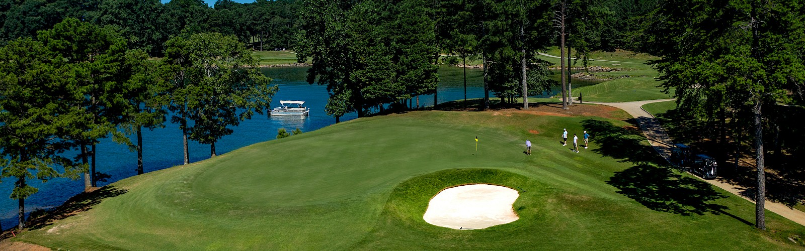 A scenic golf course with players, a water body, trees, a sand trap, and a building on the hill in the background.