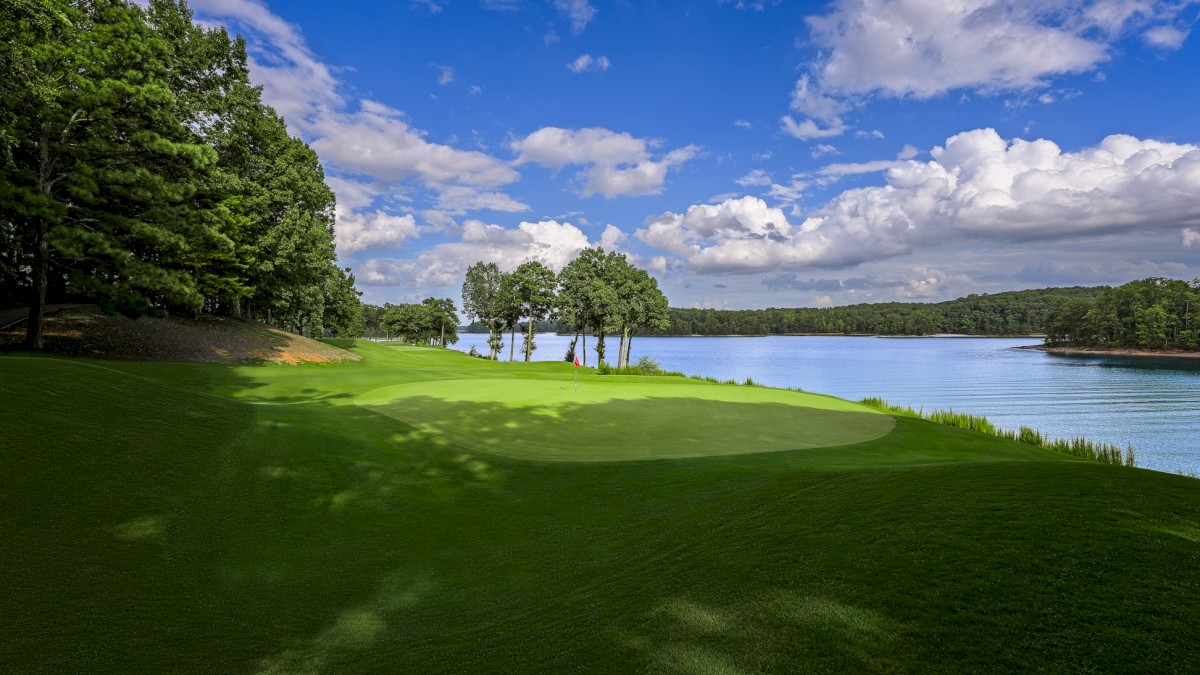 A scenic golf course beside a lake, with lush green grass, trees, and a partly cloudy sky.