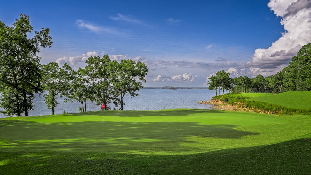 A scenic golf course near a body of water with trees, lush green grass, a flag on the hole, and a partly cloudy blue sky.