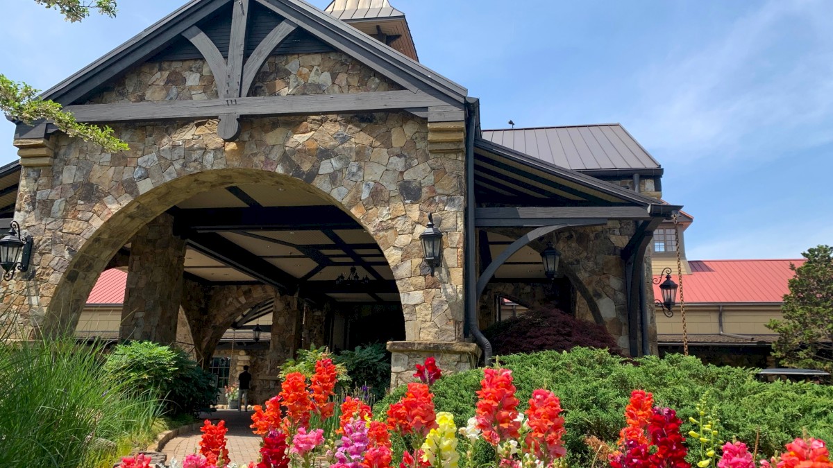A stone building with an arched entrance, surrounded by vibrant flowers and greenery, under a clear blue sky.