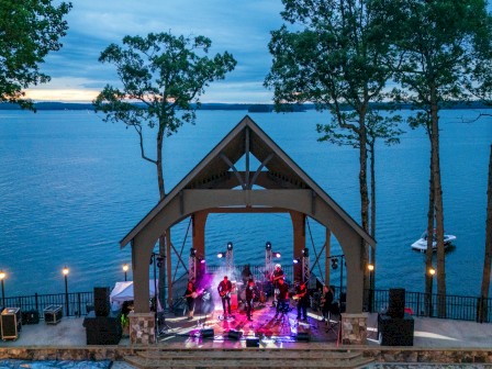 A live outdoor concert is taking place on a stage by a lake at dusk, with trees surrounding the performance area and a boat docked nearby.