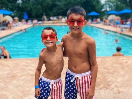 Two boys in patriotic swim trunks and red star-shaped sunglasses smiling at the swimming pool.