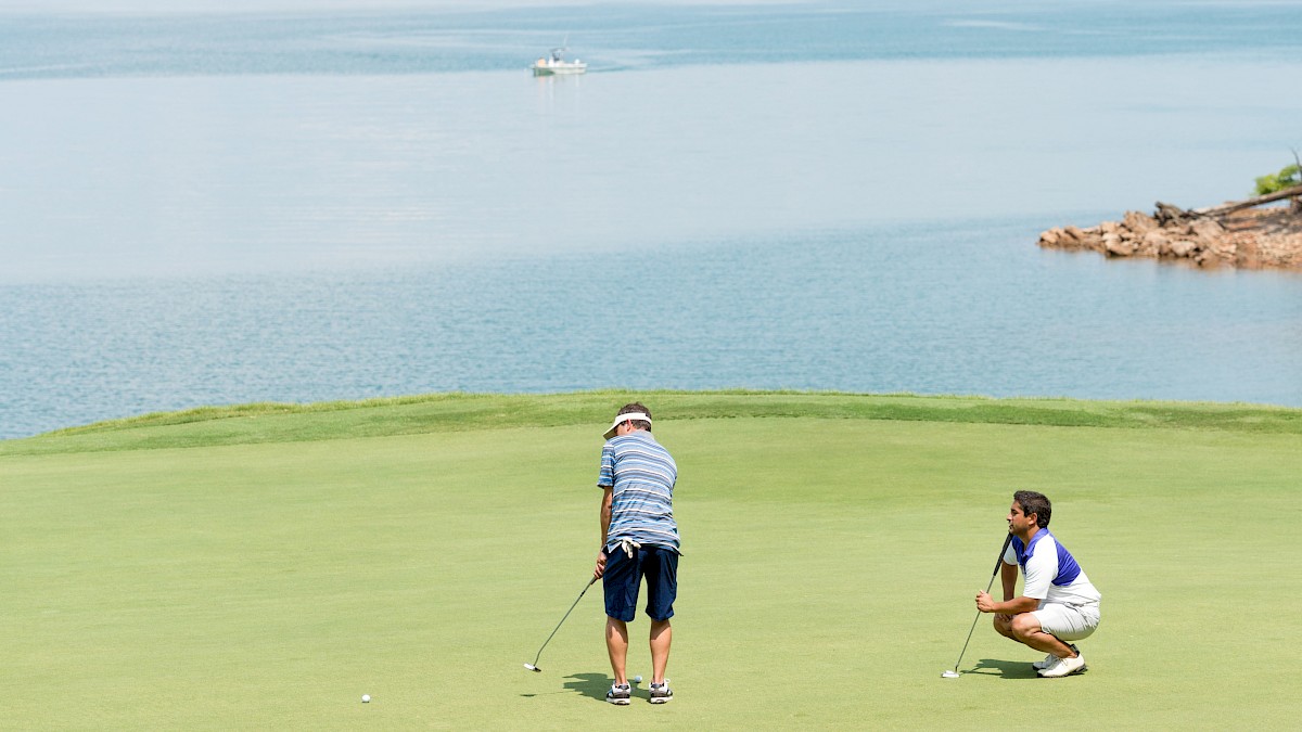 Two men play golf on a course next to a large body of water with islands, trees, and a boat in the background.