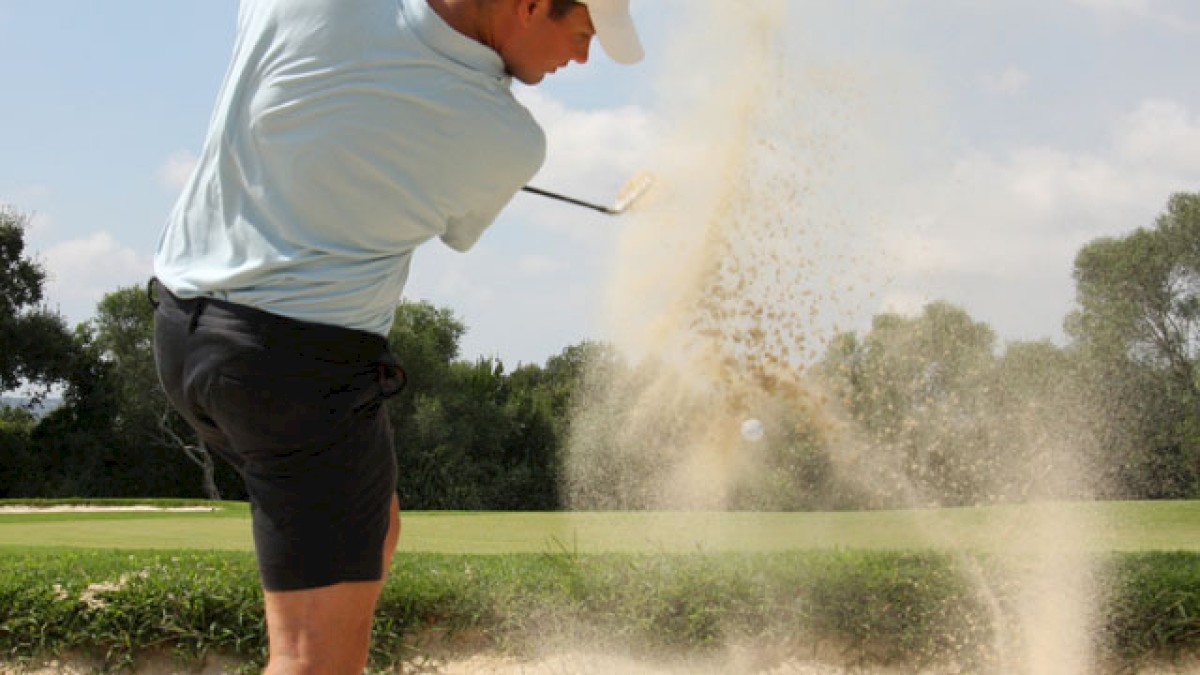 A golfer hits a shot from a sand trap, causing a sand explosion on the golf course under partly cloudy skies.