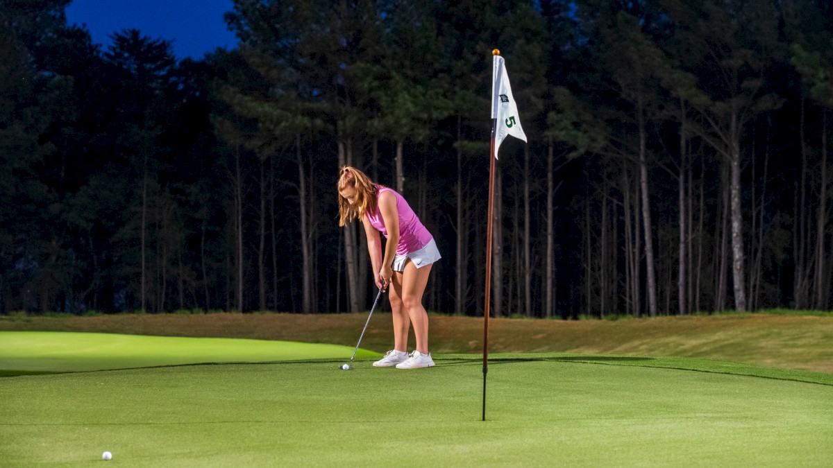 A woman in a pink top and white shorts prepares to putt on a golf green at dusk, with trees in the background.