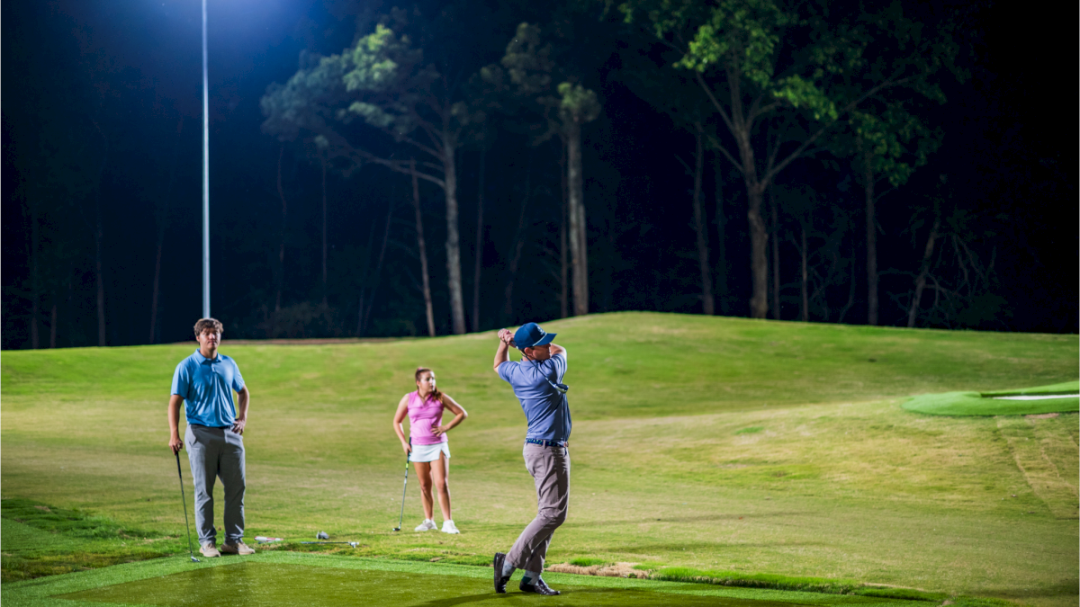Three people are playing golf on a course at night, with a golfer swinging and two others watching under bright lights.