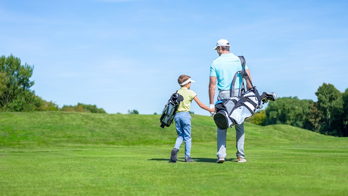 A man and a boy hold hands while walking on a golf course, both carrying golf bags, under a bright blue sky.