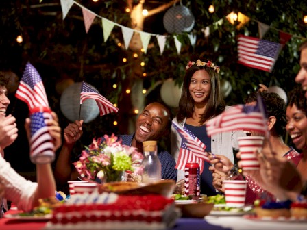 A diverse group of people celebrate with American flags, flowers, food, and festive decorations in an outdoor setting at night.