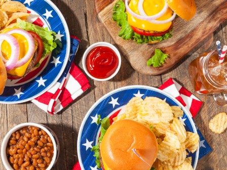 American-style burgers, chips, watermelon, baked corn, baked beans, and condiments on a rustic wooden table.
