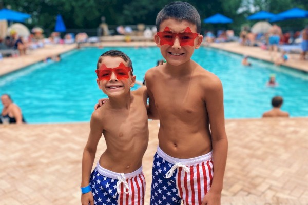 Two boys in star-spangled swim trunks and red star sunglasses pose happily at a pool on a cloudy day.
