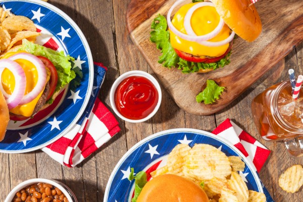A patriotic-themed picnic with burgers, chips, corn on the cob, watermelon, baked beans, and drinks on a rustic wooden table.