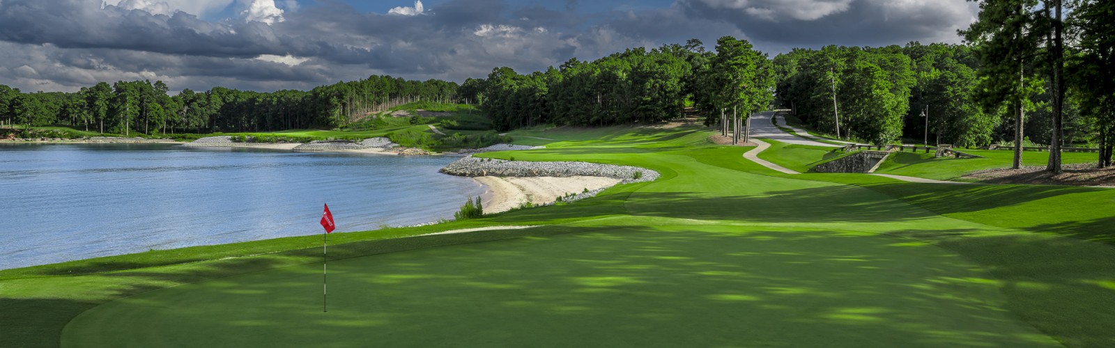 A scenic golf course near a body of water with lush green fairways, trees, sand traps, and a partly cloudy sky.