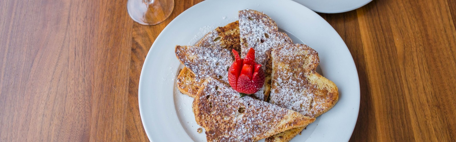 A wooden table with a morning feast: plates of crepes dusted with powdered sugar and berries, toast, a glass of orange juice, and a fancy dessert drink.
