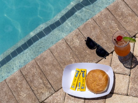 A poolside scene with sunglasses, a drink, a bread roll, and butter packets on a tiled deck.
