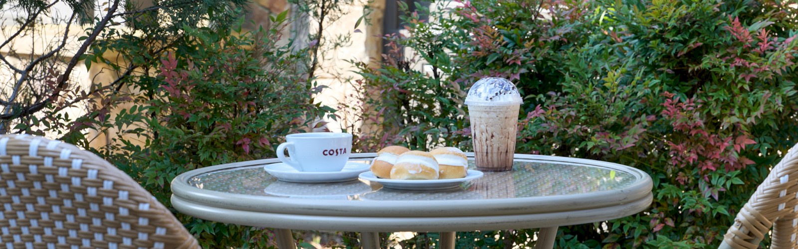 A cozy outdoor cafe table with two cups, a pastry, and a milkshake on a round table, surrounded by greenery and wicker chairs.