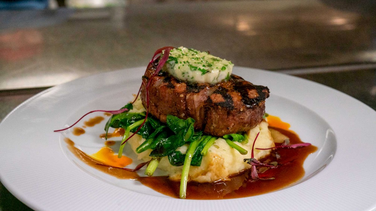 The image shows a plated meal with a seared steak topped with herb butter, served over mashed potatoes and garnished with greens and sauce.