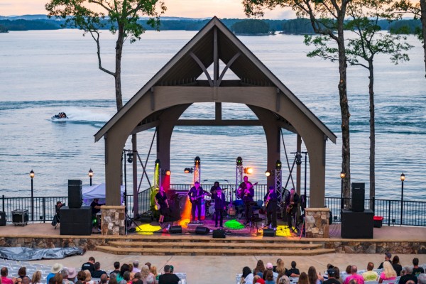 An outdoor concert by a band playing on a stage near a lake at sunset, with a large audience watching.