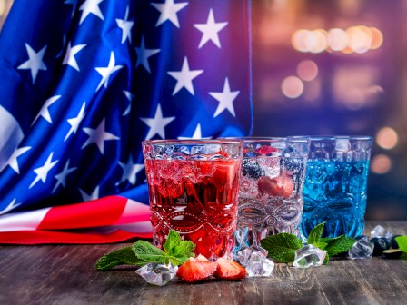 Colorful patriotic drinks with ice, strawberries, mint, set against an American flag background.