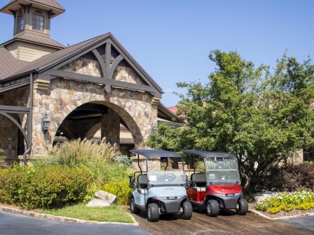 Two golf carts are parked in front of a stone building with greenery and a blue sky overhead.