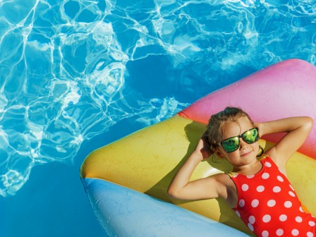 A girl in sunglasses relaxes on a colorful float in a bright blue swimming pool on a sunny day.