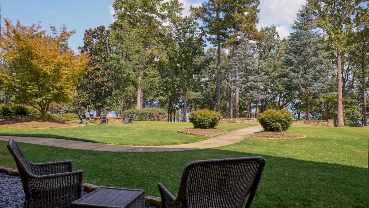 A peaceful backyard with chairs, a stone fire pit, and a lush green lawn surrounded by tall trees under a partly cloudy sky.
