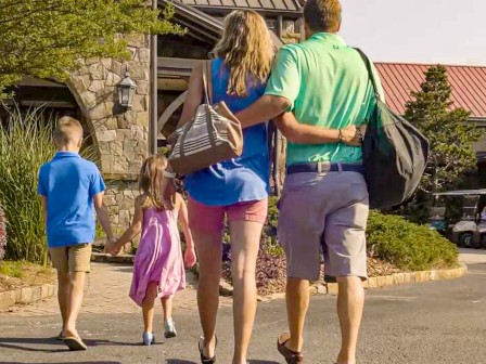 A family of five walks together outside a stone building on a sunny day, carrying bags and holding hands.
