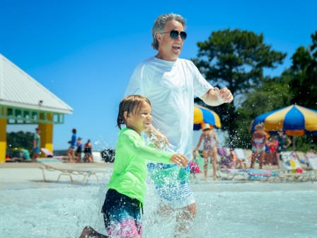 An older man and a young girl are happily playing in the water at a lively beach on a sunny day.