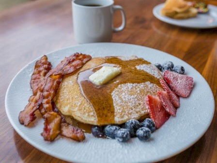 A plate of pancakes topped with butter, surrounded by bacon, strawberries, blueberries, with a coffee mug in the background.