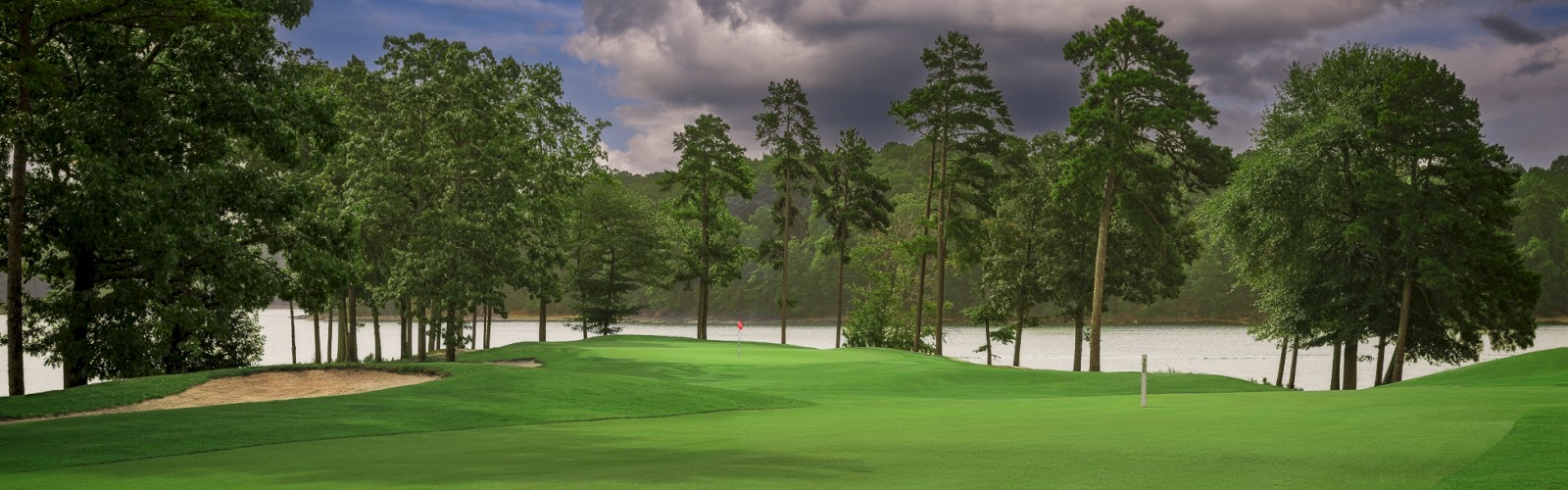 A scenic golf course with lush green fairways, tall trees, a water body in the background, and a cloudy sky overhead.